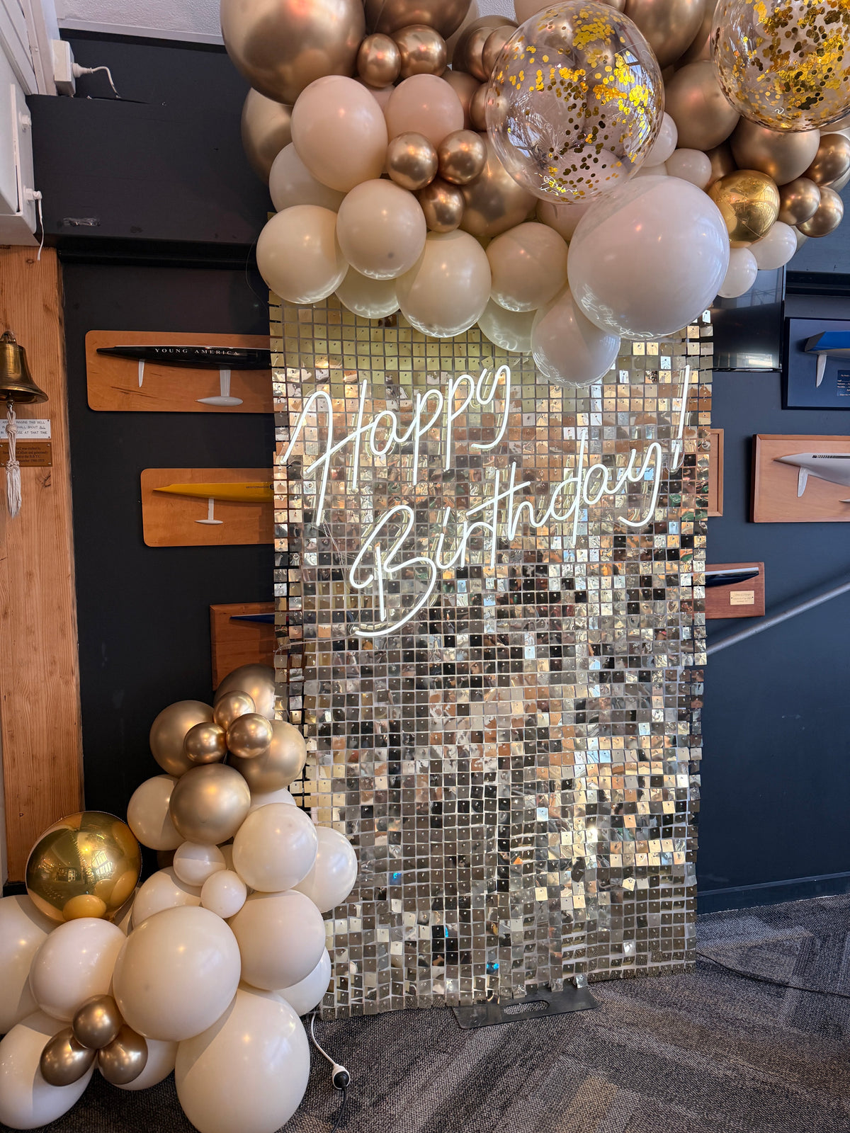 White-gold shimmer wall with Happy Birthday neon and cream-and-gold balloon garland