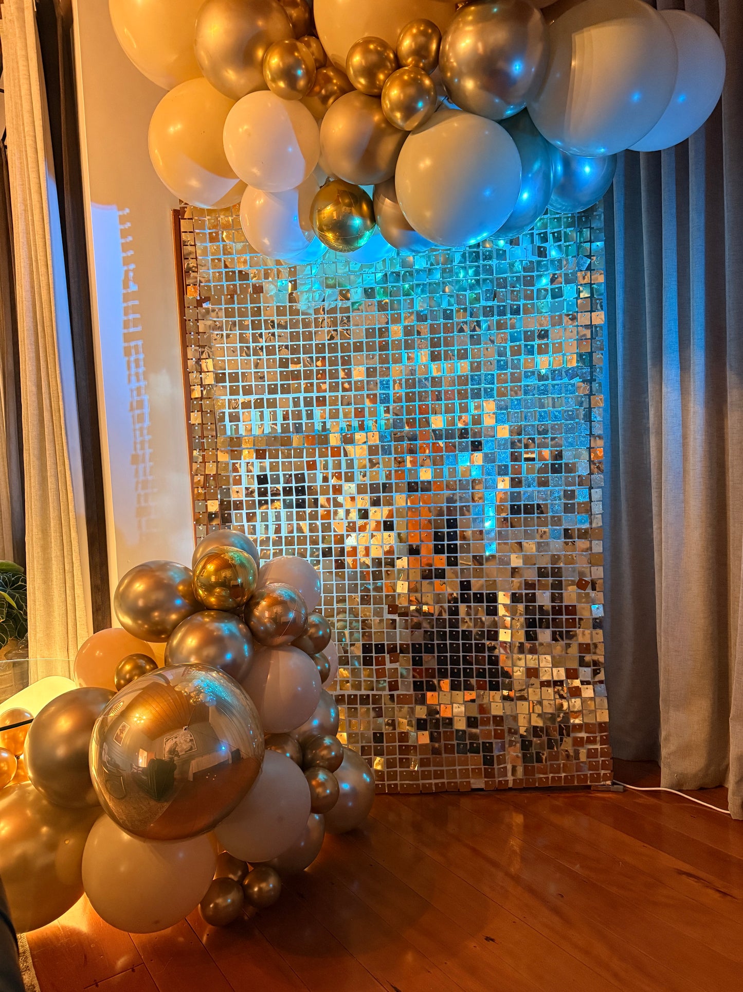 White-gold shimmer wall with cream, gold and clear confetti balloons under blue event lighting