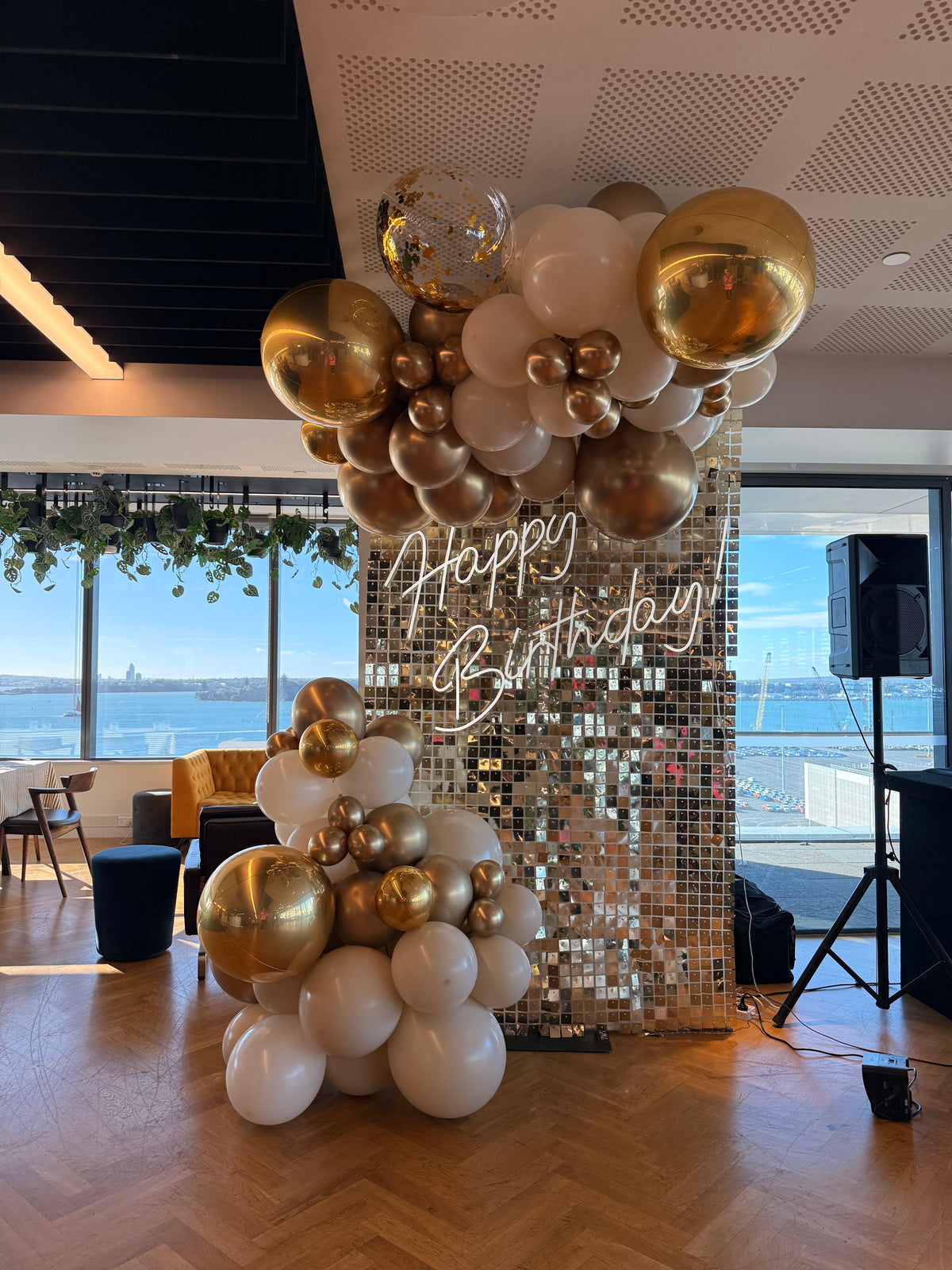 White-gold shimmer wall with Happy Birthday neon and cream-and-gold balloons at an Auckland waterfront venue